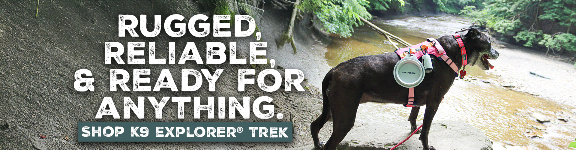 A black dog wearing pink collar, leash, and harness with grey bowl and light/waste bag combo hanging from the harness stands on a rocky ledge overlooking a river. The background features lush green trees and the river flowing below. Text on the left side of the image reads, 'RUGGED, RELIABLE, & READY FOR ANYTHING. SHOP K9 EXPLORER® TREK.'